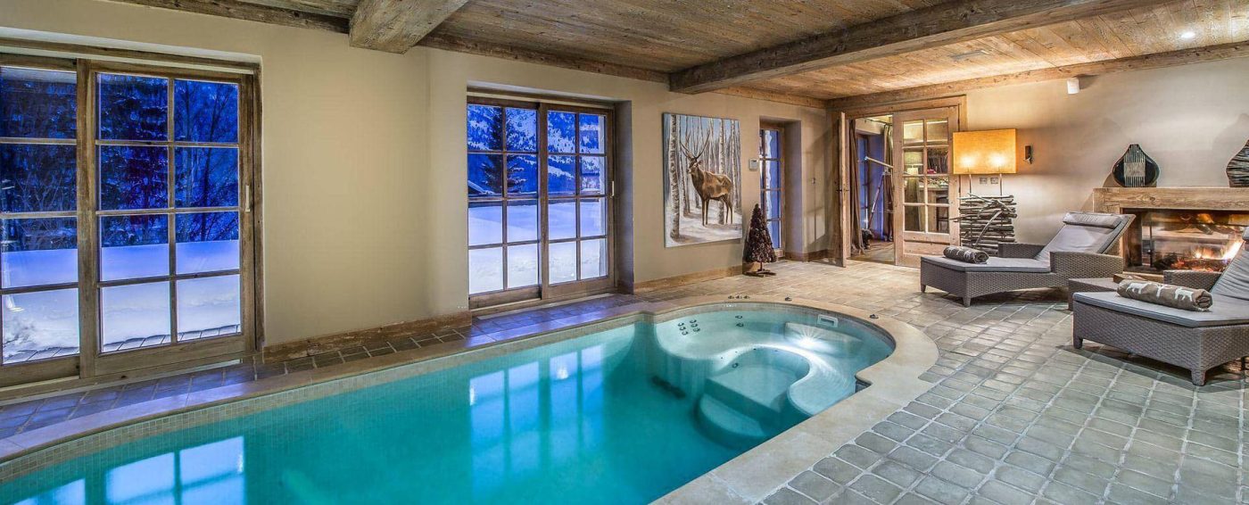 Indoor pool room with tiled floor and wood-beamed ceiling, lounge chairs by a fireplace, large windows reveal snowy outdoors—perfect for relaxing during Ski & Winter Holidays. Evening light creates a cozy, inviting atmosphere with deer wall art.